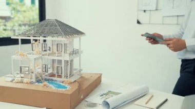 Profesyonel yakışıklı Asyalı proje yöneticisi ev modelini kontrol etmek için tablet kullanırken ev modelinin yapısını dikkatlice incelerken arkasında da planlar var. Yaratıcı tasarım. Tertemiz..