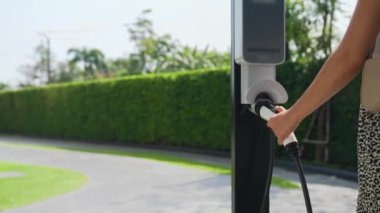 Yavaş çekim kadın, yeşil sürdürülebilir şehir parkı bahçesinde şarj olan EV elektrikli arabasıyla geziyor. Sürdürülebilir kentsel yaşam tarzında, yeniden şarj edilebilir elektrikli araç iç organlarıyla alışverişe gidiyor.