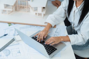 Profesyonel Asyalı erkek mimar ev modelinin uzunluğunu ölçmek için cetvel kullanırken, beyaz tenli genç meslektaşı ev modeliyle toplantı masasındaki verileri analiz etmek için dizüstü bilgisayar kullanıyor. Tertemiz..