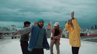 Çok kültürlü veya çok kültürlü hipster grupları şık ve renkli giysiler giyerek çatıda şehir manzaralı ve gökyüzü kazıyıcılarıyla dans ederler. Canlı sokak dansı konsepti. Açık hava sporu 2024 hiphop..