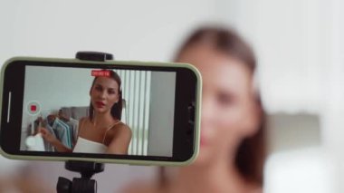 Genç sosyal medyum kadın moda videosu yapıyor. Blogcu kameraya selfie çubuğu ve ışık halkası kullanarak gülümserken, aynı zamanda ikna edici çevrimiçi kıyafetlerin izleyicilere veya takipçilere vlog satmasını sağlıyor. Düzenle