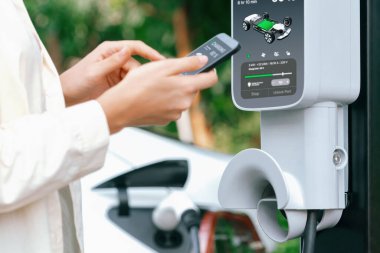 Genç bir kadın EV otomobil şarj istasyonu Green City Park 'ta elektrik ödemek için akıllı telefonu kullanıyor. Modern çevre ve EV araçlı sürdürülebilir şehir yaşam tarzı. Yararlı