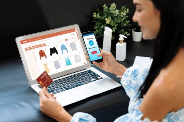 Genç bir kadın dizüstü bilgisayarla ürün sipariş eder ya da satın alır ve akıllı telefondan işlem ödemesi yapar. İnternet bankacılığı vasıtasıyla internet üzerinden kredi kartıyla online alışveriş hayatı. Blithe