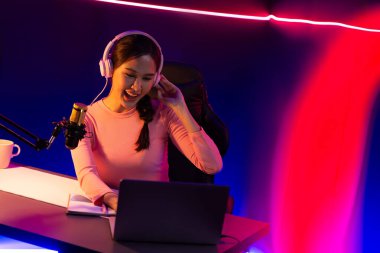 Geceleri kırmızı çerçevede podcast konuşması yaparak pembe neon ışıklı modern stüdyoda sunum için dizüstü bilgisayar kullanarak gülümseyen yaratıcı, güzel Asyalı bir etkileyiciydi. Taktik.