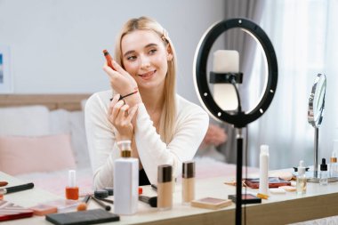 Sosyal medya için güzellik ve kozmetik video içeriği hazırlayan genç bir kadın. Seyirciye veya takipçiye nasıl sıvı ruj sürüleceğini gösterirken kamera ve ışık halkası kullanan bir güzellik blogcusu. Blithe