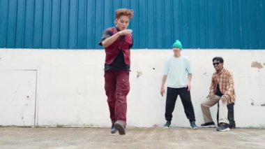 Genç Asyalı dansçı hip hop tarzında dans ediyor ve çok kültürlü arkadaşları duvarın arkasında tezahürat yapıyor. Çekici yakışıklı adam mavi arka planda kollarını uzatıyor. Açık hava sporu 2024 hiphop..