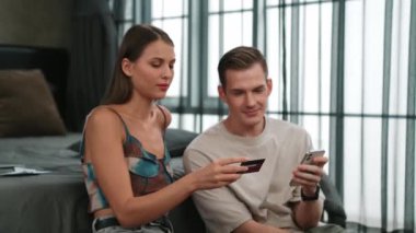 Genç çift, kredi kartıyla ödemek için internet üzerinden ödeme uygulaması ve dijital cüzdan kullanarak yatak odasında oturuyor. Mobil internet üzerinden alışveriş ve modern alımlar. Düzenle