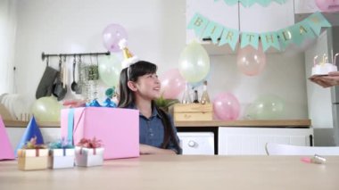 Kafkasyalı anne partide Asyalı kızların doğum gününü renkli pastayla kutladı. Mutlu anne pastayı tutarken ve odaya girerken kızı da modern mutfakta doğum günü pastasını açıyor. Pedagoji.