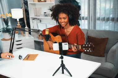 Genç Afrikalı Amerikalı müzisyenin sunucu kanalı gitar çalıyor, şarkı söylüyor, stüdyoda akıllı telefondan yayın yapıyor. Ekipman kulaklıkları dekorasyonu ve mikrofon kaydı. Tatlandırıcı.