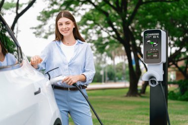 Açık yeşil şehir parkı manzaralı EV şarj istasyonundan genç kadın EV elektrikli araçları şarj ediyor. Çevre dostu şehir ulaşımı ve çevre dostu EV araba seyahati. Yükselt