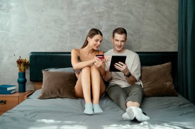 Genç çift, kredi kartıyla ödemek için internet üzerinden ödeme uygulaması ve dijital cüzdan kullanarak yatak odasında oturuyor. Mobil internet üzerinden alışveriş ve modern alımlar. Açılıyor