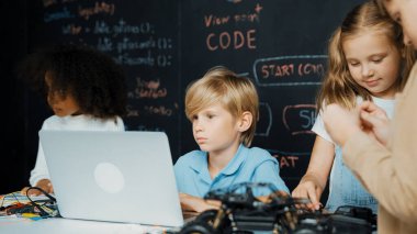 Çocuğun dizüstü bilgisayar mühendisliği kodunu ve yazım programını kullanırken bir grup zeki öğrencinin etrafı STEM teknoloji sınıfında arkadaşlarıyla çevriliyken karatahtaya oturması. Etkinlik.