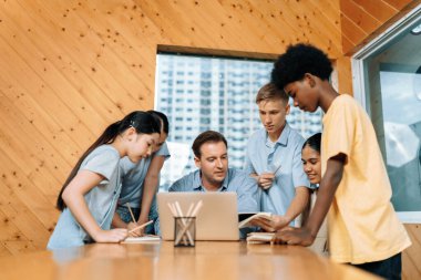 Öğretmen oturuyor ve sınıftaki çocuklara dizüstü bilgisayar kullanarak ders veriyor, çocuklarla defterdeki içerik hakkında konuşuyor. Konunun ne olduğunu soran öğrenciler onlara öğretiyor. Öğretim.
