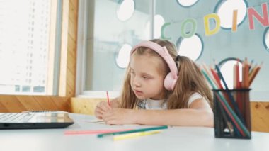 Öğretmenini dinlerken sınıfta ya da internette öğrenim gören tatlı bir kız. Mutlu öğrenci yazıları, çizimleri, bilgisayar ve renkli kalemlerle kulaklık takarak kağıt üzerinde çalışması. Odaklan. Etkinlik.