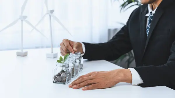 stock image Businessman put coin to money saving glass jar at his office as sustainable money growth investment or eco-subsidize. Green corporate promot and invest in environmental awareness. Gyre