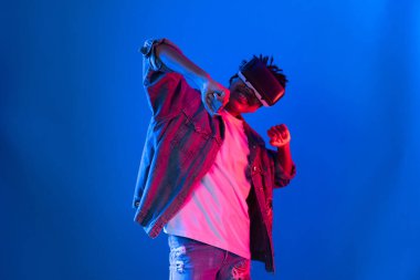 Gülümseyen Afrikalı genç Amerikalı sanal gerçekliği dijital fütüristik teknolojiye bağlayan pembe mavi neon duvarda hologram oyunlarıyla VR dansına bakıyor. Entrika.