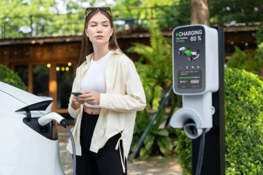 İlkbahar bahçesindeki açık kafede çalışan EV elektrikli araba şarjı, yeşil şehir sürdürülebilirliği ve çevre dostu EV otomobili için genç bir kadınla sürdürülebilir şehir seyahati elektrik ödüyor. Yararlı