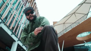 Hip hop tarzında düşük açılı hipster pozu alışveriş merkezi veya bina manzarası ile çevrilidir. Kameraya bakarken Asyalı hip hop koreografı break dans yapıyor. Açık hava sporu 2024 hiphop..