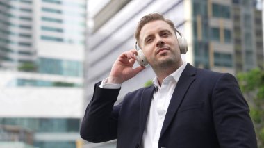 Erkek liderin etrafını sarın kulaklığı dinleyin ve neşeli ve motive edici bir şekilde rahatlatıcı müzikle dans edin. Becerikli proje yöneticisi şehir merkezinde akıllı telefon kullanırken rahat şarkılar dinlemeyi sever. Urbane.