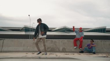 Yakışıklı break dansçı, Asyalı arkadaşının tezahürat yaptığı serbest stil hareketinden hoşlanır. Hippi kulaklıktan müzik dinler ve kameraya bakar. Sokak dansı. Açık hava sporu 2024. Yumuşak bir şekilde..