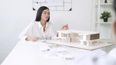 Yetenekli, genç, güzel mimar mühendisi ev modeli, taslak ve mimari ekipmanlarla toplantı masasında ev modeli hakkında bir harita tutan yakışıklı bir iş arkadaşıyla görüşüyor. Tertemiz..