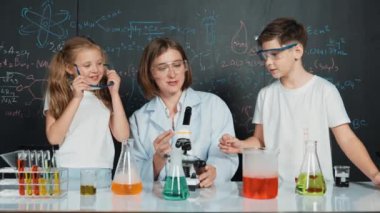 Öğretmen tavsiye verirken beyaz çocuk kimyasal sıvıyı karıştırıyor. Profesyonel eğitmen labaratuar kıyafeti giymiş, renkli solüsyonla dolu deney kabıyla sofrada çeşitli öğrencileri arıyor. Etkinlik.