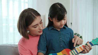 Kafkasyalı bir anne evde akustik müzik öğretirken ukulele çalan mutlu kız. Sevimli çocuk enstrüman öğreniyor. Kafkasyalı mutlu anne ve kız birlikte vakit geçiriyorlar. Pedagoji.