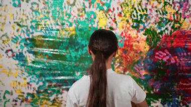 Genç ve güzel bir kızın arka görünüşü renkli duvarı boyamak için elini kaldırır. Çekici bir lise öğrencisi renk paletini tutarken duvarı el iziyle boyuyor. Yaratıcı aktivite. Öğretim