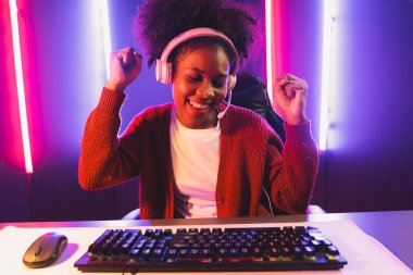 Oyun flama, Afrikalı kız Esport yetenekli takımıyla online kavga ediyor. Neon renkli aydınlatma odasında kulaklık takıyor. Rakiplerini kazanmak için strateji planlayan diğer oyuncularla konuşmak. Tatlandırıcı.