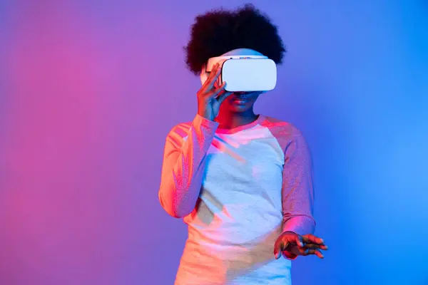 stock image Young African wearing VR glasses through metaverse to select 3D hologram data application isolated on blue pink neon lighting gradient background in technology digital innovation graphic. Contrivance.