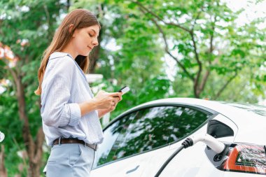 Ulusal Park veya Yaz Ormanı 'ndaki tatil gezisi sırasında EV şarj istasyonundan elektrik aküsü almak için akıllı telefon internet bankacılık uygulaması kullanan genç bir kadın. Yükselt