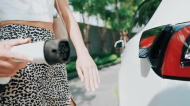 Yavaş çekim kadın, yeşil sürdürülebilir şehir parkı bahçesinde şarj olan EV elektrikli arabasıyla geziyor. Sürdürülebilir kentsel yaşam tarzında, yeniden şarj edilebilir elektrikli araç iç organlarıyla alışverişe gidiyor.