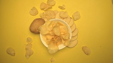 Patates cipsinin yavaş çekimde sarı arka planlı beyaz kaseye düşüşü. Kızarmış patateslerin üst tarafı patatesle dolu kaseye düşer. Ağır çekim. Makrografi. Kapatın. Geliştirilebilir.