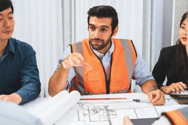 Mimari proje üzerinde birlikte çalışan çeşitli inşaat mühendisi ve müvekkil grupları, inşaat planını gözden geçirip toplantı masasında plan oluşturuyorlar. Tedbirli