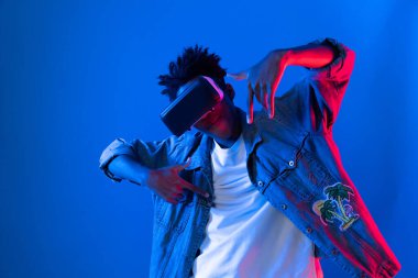 Gülümseyen Afrikalı genç Amerikalı sanal gerçekliği dijital fütüristik teknolojiye bağlayan pembe mavi neon duvarda hologram oyunlarıyla VR dansına bakıyor. Entrika.