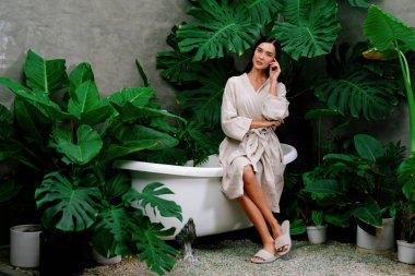 Modern otelde küveti olan tropikal ve egzotik bir spa bahçesi ya da yemyeşil yeşillik arka planıyla çevrili rahat ve sağlıklı yaşam tarzının tadını çıkaran bornozlu bir kadınla tatil köyü. Blithe