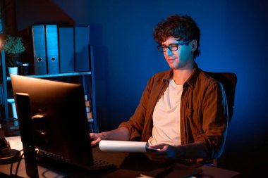 Genç işadamı, Neon Light Modern Ofis 'teki yeni proje surround çalışma masası için kağıt üzerinde tasarım düşünüyor. E-posta ile hediye göndermeden önce not defterine yeni bir iş düşünüyorum. Toplayıcı.