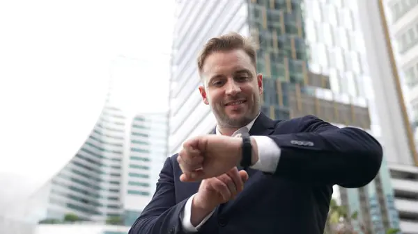 Low angle camera of businessman looking at watch to remind about time. Smart caucasian businessman checking time for being on time in important appointment or meeting with modern skyscraper. Urbane.