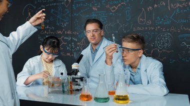 Yaratıcı öğretmen masadaki liseli çocukla mikroskop ve renkli sıvıyla test tüpüyle konuşurken kimyayı tahtaya doğrultuyor. BTMM sınıfında laboratuvar önlüğü giyen genç bir öğrenci. Öğretim.