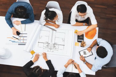 Mimari proje üzerinde birlikte çalışan, inşaat planını gözden geçiren ve toplantı masasında plan oluşturan çeşitli inşaat mühendisi ve müşteri gruplarından oluşan üst düzey bir pankart. Tedbirli