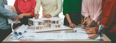 Profesyonel mimar takımının ev rengi seçimi hakkında beyin fırtınası yaparken, mimar da plan ve ev modelinin olduğu masadaki taslak belgeye yazı yazıyor. Değiştirilmiş.