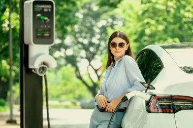 Yolculuk sırasında doğal orman ya da milli parktaki elektrikli araba için şarj olan genç bir kadın. Tatil ve tatil boyunca çevre dostu seyahat. Yükselt