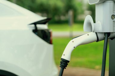 Açık yeşil şehir parkı manzarasındaki EV şarj istasyonundan EV elektrikli araç şarj oluyor. Çevre dostu EV araba yolculuğuyla doğal koruma. Yükselt