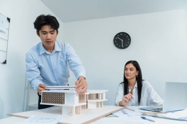 Profesyonel Asyalı erkek mimar ev modelinin uzunluğunu ölçmek için cetvel kullanırken, beyaz tenli genç meslektaşı ev modeliyle toplantı masasındaki verileri analiz etmek için dizüstü bilgisayar kullanıyor. Tertemiz..