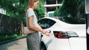 Şehir merkezindeki EV şarj istasyonunda elektrikli araba aküsünü yavaş çekimde şarj eden kadın elektrikli aracın iç organları için sürdürülebilir yeşil ve yenilenebilir temiz enerji yaşam tarzına sahip.