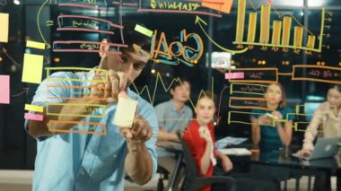 Professional businessman putting sticky notes on glass wall while explain start up project. Skilled project manager pointing at business plan while sharing marketing idea to investor. Tracery