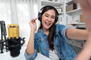 Sunucu kanalı genç ve güzel Asyalı kız akıllı telefonuyla selfie çekiyor. Modern stüdyo ofis konseptinde kulaklık takıyor. Sahne arkası yayıncısı sosyal medyada canlı yayın yapıyor. Taktik.