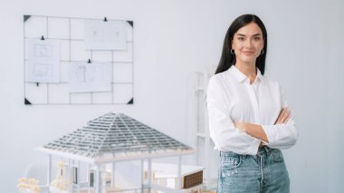 Genç, güzel, profesyonel mimar mühendisin portresini yakın plan ile birlikte beyaz modern ofiste ev modeliyle güven içinde pozlar veriyor. Yaratıcı yaşam ve tasarım. Tertemiz..