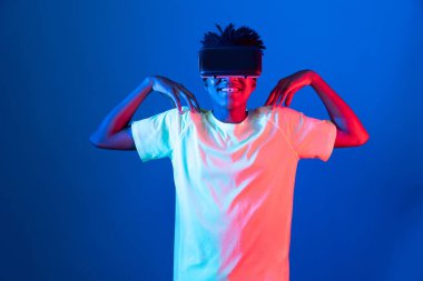 Genç Afro-Amerikalı sporcu VR 'ı giyip kollarını üst tarafa uzatarak mavi neon ışıklı duvara poz vererek metaevren dünyasındaki dijital fütüristik teknolojiyi sanal gerçekliğe bağlıyor. Entrika.
