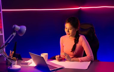 Geceleri sosyal medyada canlı yayında pozitif podcastla konuşan gülümseyen genç, güzel Asyalı etkileyici pembe neon ışıklı modern stüdyoda sunum için dizüstü bilgisayar kullanıyor. Taktik.